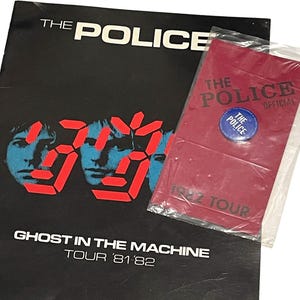 May include: A black The Police Ghost in the Machine tour program from 1981-82 featuring red digital-style numbers and band member portraits. Included is a red The Police Official 1982 Tour envelope containing a small blue circular pin badge.