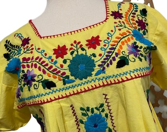 May include: Yellow embroidered cotton dress with colorful floral designs. The dress has a square neckline and short sleeves.