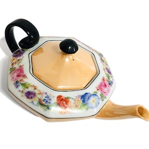 May include: A white porcelain teapot with a floral design and a black handle. The teapot has a lid with a black knob and a floral design. The teapot is on a white background.