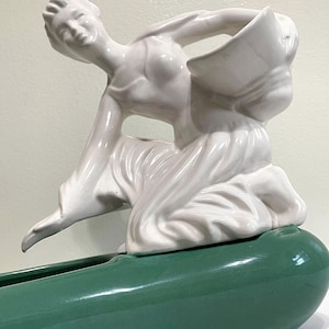 May include: A white ceramic figurine of a woman holding a bowl, sitting on a green ceramic planter. The woman is in a flowing pose, and the planter is shaped like a long, narrow boat.