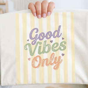 Puede incluir: Camiseta color crema con un fondo a rayas amarillo pastel. Las palabras "Good Vibes Only" están escritas en una fuente caprichosa con letras moradas, verdes y naranjas. Pequeños corazones adornan el texto.