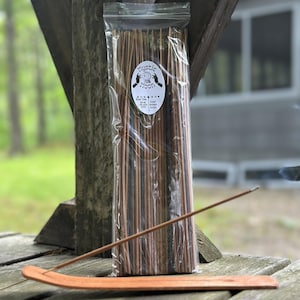 May include: A clear plastic bag containing a bundle of brown incense sticks with a white oval label reading Mystic Moon, featuring a graphic of hands and a crystal ball. A single incense stick rests on a curved wooden incense burner tray.