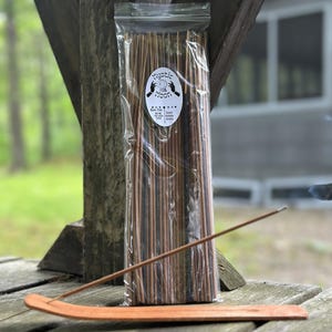 May include: A clear plastic bag containing a bundle of brown incense sticks with a white oval label featuring the text Mystic Moon and a graphic of hands holding a crystal ball. A single incense stick rests on a curved wooden incense holder.