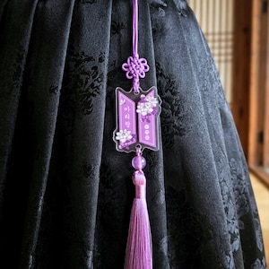 Puede incluir: Un dije de acr&iacute;lico morado con ilustraciones florales blancas y texto en coreano, suspendido por un cord&oacute;n morado a juego con un nudo decorativo y una borla larga y sedosa de color morado, sujeto a una prenda de tela negra con un broche de metal plateado.