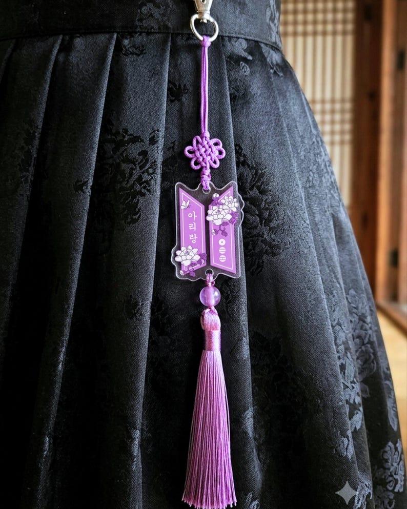 Puede incluir: Un dije de acr&iacute;lico morado con ilustraciones florales blancas y texto en coreano, suspendido por un cord&oacute;n morado a juego con un nudo decorativo y una borla larga y sedosa de color morado, sujeto a una prenda de tela negra con un broche de metal plateado.