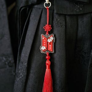 Puede incluir: Un llavero de acr&iacute;lico rojo y blanco con un cierre de mosquet&oacute;n plateado, un nudo decorativo rojo y una borla roja larga. El dije muestra detalles florales blancos y el texto coreano "아리랑" sobre un fondo rectangular rojo.