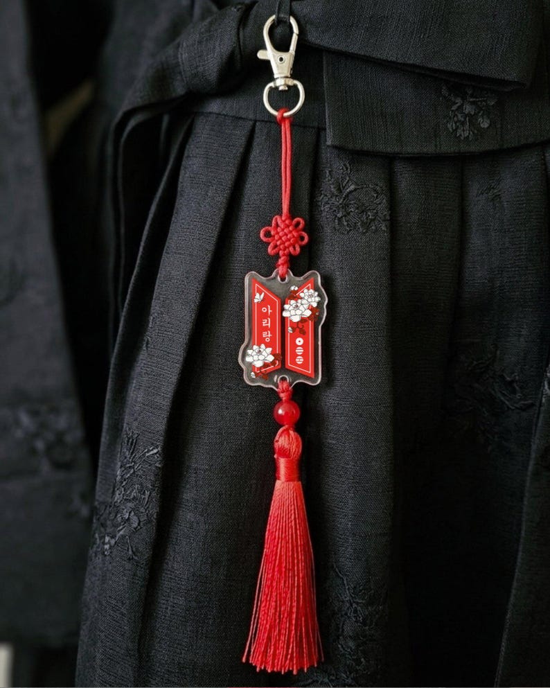 Puede incluir: Un llavero de acr&iacute;lico rojo y blanco con un cierre de mosquet&oacute;n plateado, un nudo decorativo rojo y una borla roja larga. El dije muestra detalles florales blancos y el texto coreano "아리랑" sobre un fondo rectangular rojo.