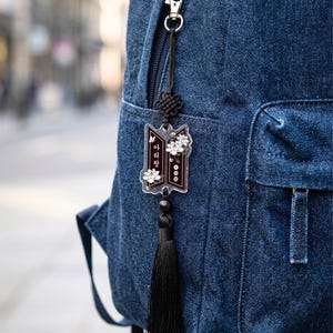 Puede incluir: Una mochila de mezclilla azul oscuro con un dije de acr&iacute;lico colgante. El dije tiene forma rectangular oscura con detalles florales blancos y texto en coreano, suspendido por un cord&oacute;n negro con un nudo decorativo y una borla de tela negra.