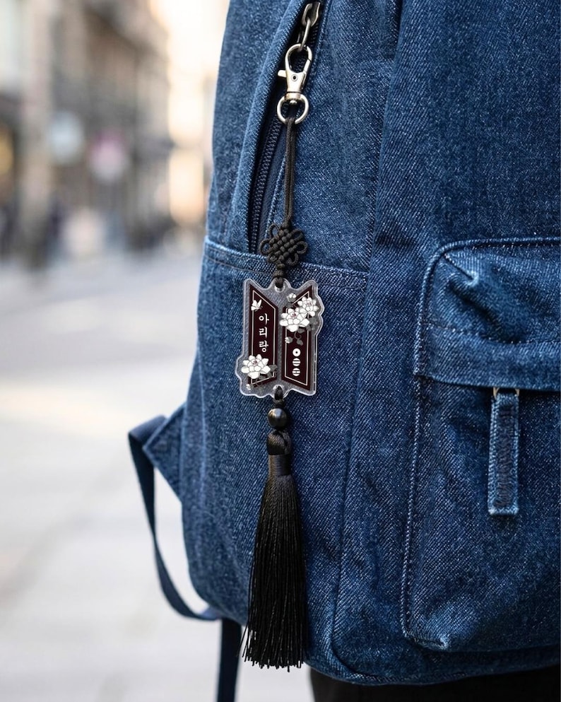 Puede incluir: Una mochila de mezclilla azul oscuro con un dije de acr&iacute;lico colgante. El dije tiene forma rectangular oscura con detalles florales blancos y texto en coreano, suspendido por un cord&oacute;n negro con un nudo decorativo y una borla de tela negra.