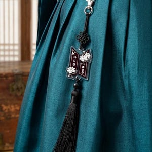 Puede incluir: Una prenda de tela plisada en color verde azulado oscuro que presenta un accesorio colgante con un cierre de metal plateado, un cord&oacute;n negro con un nudo decorativo, un dije de acr&iacute;lico transparente con flores blancas y texto coreano, y una borla de hilo negro.