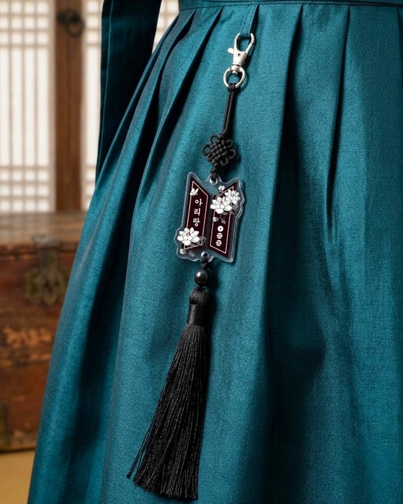 Puede incluir: Una prenda de tela plisada en color verde azulado oscuro que presenta un accesorio colgante con un cierre de metal plateado, un cord&oacute;n negro con un nudo decorativo, un dije de acr&iacute;lico transparente con flores blancas y texto coreano, y una borla de hilo negro.