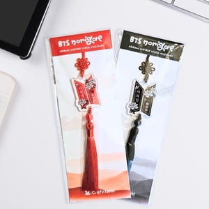 Puede incluir: Dos accesorios de borla BTS norigae inspirados en Arirang en envases de pl&aacute;stico transparente. Uno es rojo con borla y nudo rojos, el otro es negro con borla y nudo negros. Ambos cuentan con un dije con detalles florales y texto en coreano.