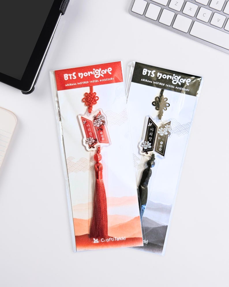 Puede incluir: Dos accesorios de borla BTS norigae inspirados en Arirang en envases de pl&aacute;stico transparente. Uno es rojo con borla y nudo rojos, el otro es negro con borla y nudo negros. Ambos cuentan con un dije con detalles florales y texto en coreano.