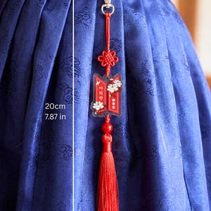 Puede incluir: Un colgante decorativo rojo con cierre de mosquet&oacute;n plateado, un nudo rojo y una borla roja. El colgante incluye una pieza de acr&iacute;lico transparente con gr&aacute;ficos florales blancos y texto en coreano. Una l&iacute;nea de medici&oacute;n indica 20 cm.