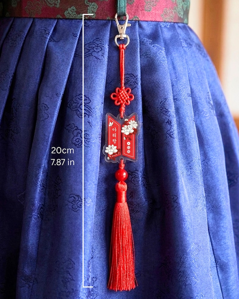 Puede incluir: Un colgante decorativo rojo con cierre de mosquet&oacute;n plateado, un nudo rojo y una borla roja. El colgante incluye una pieza de acr&iacute;lico transparente con gr&aacute;ficos florales blancos y texto en coreano. Una l&iacute;nea de medici&oacute;n indica 20 cm.
