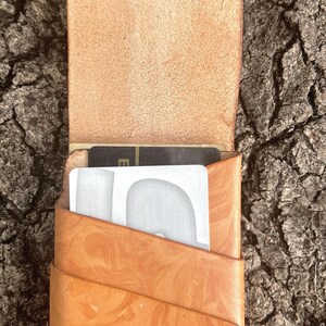 May include: A tan leather card holder with a marbled pattern and a vertical pocket design. The wallet features a flap closure and contains two visible cards, one black and one silver, tucked into the front slot with white stitching at the base.