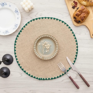 May include: A round, tan, woven placemat featuring a dark green stitched scalloped edge. Centered on the placemat is a small, round, light-coloured plate containing a delicate sprig of dried white flowers, accompanied by a silver fork and knife.