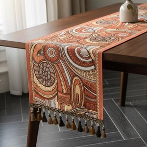 Könnte beinhalten: Dekorativer Tischläufer mit einem abstrakten Design in Orange-, Braun- und Cremetönen. Der Läufer weist ein sich wiederholendes Muster aus Wirbeln und Kreisen auf und ist mit einem braunen Rand und Quasten versehen.