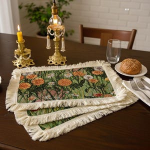 May include: A stack of three rectangular placemats with a floral design featuring orange and white flowers on a green background. The placemats have a cream-colored fringe border. The scene includes a candle, a light fixture, and a roll.