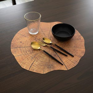 Peut inclure: Un set de table rond arborant un motif réaliste de grain de bois brun rappelant une coupe transversale de tronc d'arbre. Il est disposé avec un verre texturé transparent, un bol noir mat et des couverts dorés à manches noirs au centre.