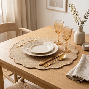 Op de afbeelding: Een gedekte tafel met een beige placemat, witte borden en gouden bestek. Twee amberkleurige wijnglazen en een kleine vaas met witte bloemen. Een gevouwen linnen servet en een kleine kom maken het geheel af.
