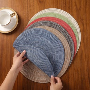 May include: A selection of circular placemats in various colours, including blue, brown, red, green, and grey. The woven placemats are arranged in a fan shape on a wooden table. A white teacup and saucer with gold trim are visible in the top left corner.