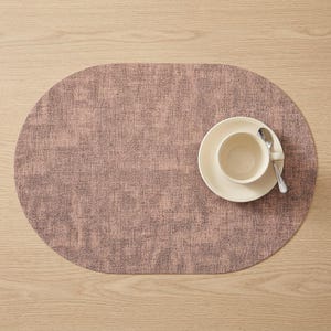 May include: An oval-shaped, mauve placemat with a textured appearance. A white teacup and saucer with a silver spoon rest on the placemat. The placemat is on a light wood surface, creating a neutral and inviting setting.
