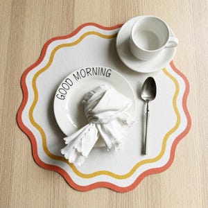 May include: A white placemat with a wavy edge, bordered by orange and yellow stripes. A white plate with "GOOD MORNING" printed on it, a folded napkin, a spoon, and a teacup and saucer are arranged on the placemat.