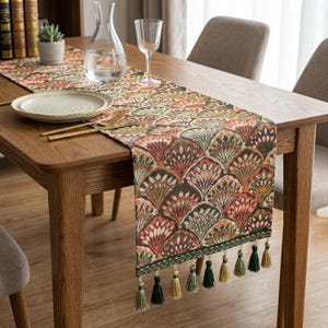 Può includere: Una tovaglia decorativa con un motivo a ventaglio ripetuto in tonalità arancioni, verdi e marroni. La tovaglia è adornata con nappe lungo il bordo inferiore. È posizionata su un tavolo di legno con un piatto, posate e un bicchiere.