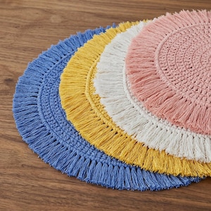 May include: A collection of colorful, round placemats with fringed edges. The placemats are in shades of blue, yellow, white, and pink. The placemats are made of a woven material and are arranged on a wooden surface.