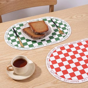 May include: A table setting with two oval placemats, one green and one red, both with a chequered design. A plate of bread, a cup of tea, and cutlery are on the table. The words "ENJOY LIFE" are printed around the edge of the placemats.