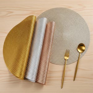 Peut inclure: Ensemble de sets de table tissés dorés, argentés et or rose, ainsi qu'un grand set de table rond gris. Une fourchette et une cuillère dorées sont posées sur une surface en bois, suggérant un cadre de repas. Les sets sont enroulés et déployés.