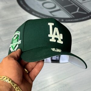 May include: A dark green baseball cap with the letters "LA" in white on the front. The cap has a flat brim with a white underside and a patch on the side. The cap is being held by a person.