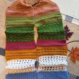 Peut inclure: Une longue écharpe en crochet rectangulaire présentant des rayures horizontales de différentes couleurs, notamment beige, rose, vert olive, vert foncé, magenta, jaune moutarde, blanc et bleu clair, avec des points pleins et ajourés.