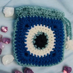 Op de afbeelding: Gehaakte tas met een evil eye-ontwerp. De vierkante tas heeft een blauw, wit en zwart oogpatroon, met een turkooizen trekkoordsluiting en witte pompons. Heldere en paarse kristallen omringen de tas.