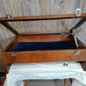 May include: A rectangular wooden display case with a hinged glass lid, featuring a deep blue velvet-lined interior. The box includes silver-toned metal latches, a support arm for the lid, and two small metal keys resting on the surface below.