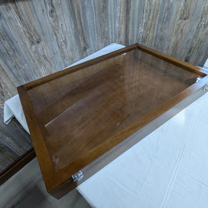 May include: A rectangular wooden display case with a clear glass top. The case has a dark brown finish and metal clasps. The case is empty and sits on a white surface.