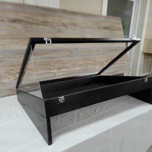 May include: A black wooden display case featuring a clear, hinged lid that opens upward. The rectangular box has a dark finish, a flat interior base, and silver-toned metal latches on the front and top edges to secure the transparent cover.
