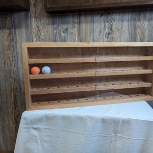 May include: A wooden display case with multiple shelves and small holes, likely for golf balls. Two golf balls, one orange and one white, are visible. The case is made of light-colored wood and has a clear front.