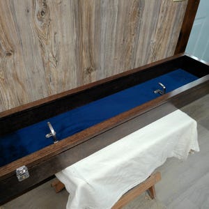 May include: A long, rectangular wooden display case with a dark brown finish and a deep blue velvet-lined interior. The case features two silver-coloured metal latches on the front edge and two silver-coloured metal hooks mounted inside the base.