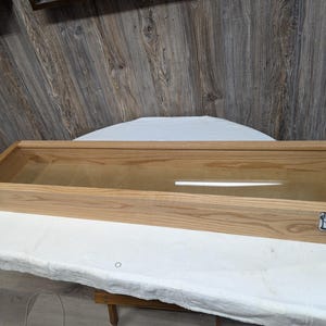 Op de afbeelding: Een rechthoekige houten vitrine met een helder glazen bovenkant. De behuizing is gemaakt van lichtgekleurd hout en heeft metalen sluitingen aan de voorkant. De behuizing is leeg en staat op een wit oppervlak.