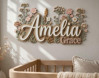 以下が含まれることがあります： 白とベージュの重ね文字で「Amelia Grace」と書かれた壁掛け看板。ピンク、白、緑、ベージュの色合いの立体的な花や葉の装飾が施されており、ニュートラルな背景に配置されています。