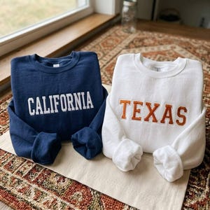 Puede incluir: Una sudadera azul marino de cuello redondo con la palabra CALIFORNIA bordada en letras de bloque blancas y una sudadera blanca de cuello redondo con la palabra TEXAS bordada en letras de bloque naranjas con un borde blanco en el frente.