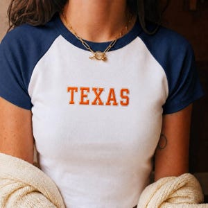 Könnte beinhalten: Ein weißes Raglan-T-Shirt mit dunkelblauen Ärmeln und Rundhalsausschnitt. Das Wort TEXAS ist in orangefarbenen Blockbuchstaben mit weißer Umrandung auf die Brust gestickt. Eine goldfarbene Kette mit Knebelverschluss wird um den Hals getragen.