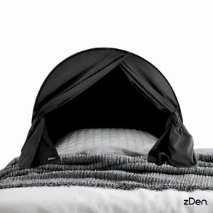 May include: A black bed canopy, draped over a pillow and blanket. The canopy has a curved top and is tied at the sides. The blanket is gray and textured. The brand name "zDen" is visible.