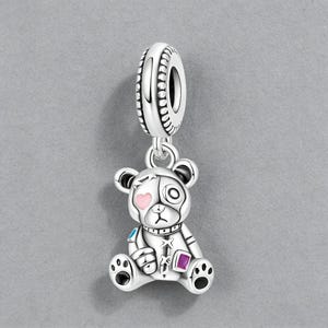 Könnte beinhalten: Ein silberner Charm-Anhänger mit einem Roboter-Teddybär-Design. Der Bär hat ein rosa Herz, ein lila Quadrat und blaue Akzente. Der Charm-Anhänger hängt an einer dekorativen Schlaufe, ideal für ein Armband oder eine Halskette.