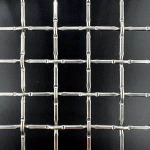May include: Close-up of a metal grid with a square pattern. The grid is made of silver-colored metal and has a repeating pattern of intersecting lines. The background is black, providing contrast to the metallic grid.