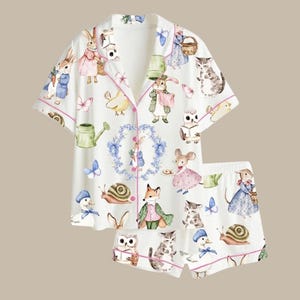 May include: White pajama set with a whimsical print of rabbits, cats, ducks, and other woodland creatures. The short-sleeved top and shorts are trimmed with pink piping. The illustrations include butterflies, watering cans, and floral accents.
