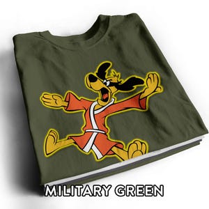 May include: A military green t-shirt featuring a graphic of a yellow cartoon dog wearing a red robe and a black eye mask, depicted in a dynamic pose with arms outstretched. The text MILITARY GREEN is printed in white at the bottom of the image.