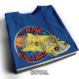 Op de afbeelding: Een koningsblauw t-shirt met een ronde afbeelding van een gele schoolbus met vleugels en raketmotoren. De tekst "The Magic School Bus" staat in het rood rond de cirkel gedrukt. Passagiers zijn zichtbaar in de ramen en "WAHOO" staat op het bord van de bus.
